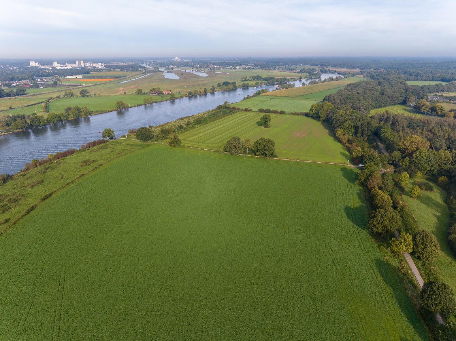 Luchtfoto van een groot groen grasland grenzend rechts aan de Maas.